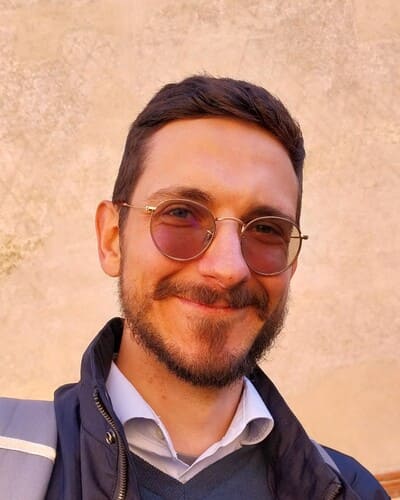 Professional headshot of a smiling young man with short brown hair, a well-groomed beard, wearing round metal-framed glasses, a light collared shirt, dark sweater, and jacket, against a neutral textured background in warm natural light